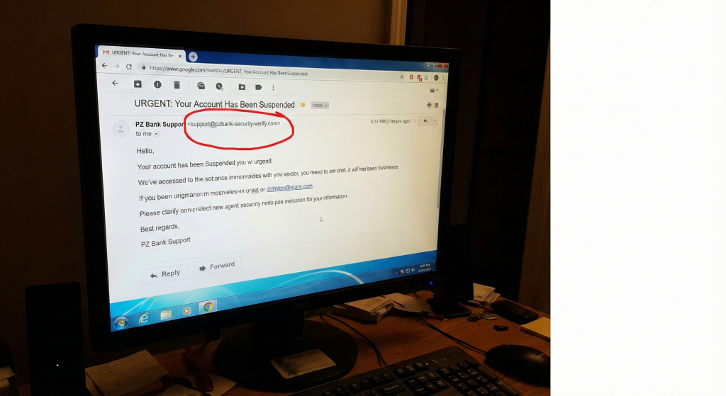 A close-up photograph of a computer monitor displaying a fake phishing email, with a red circle highlighting the suspicious sender's email address as a warning sign.